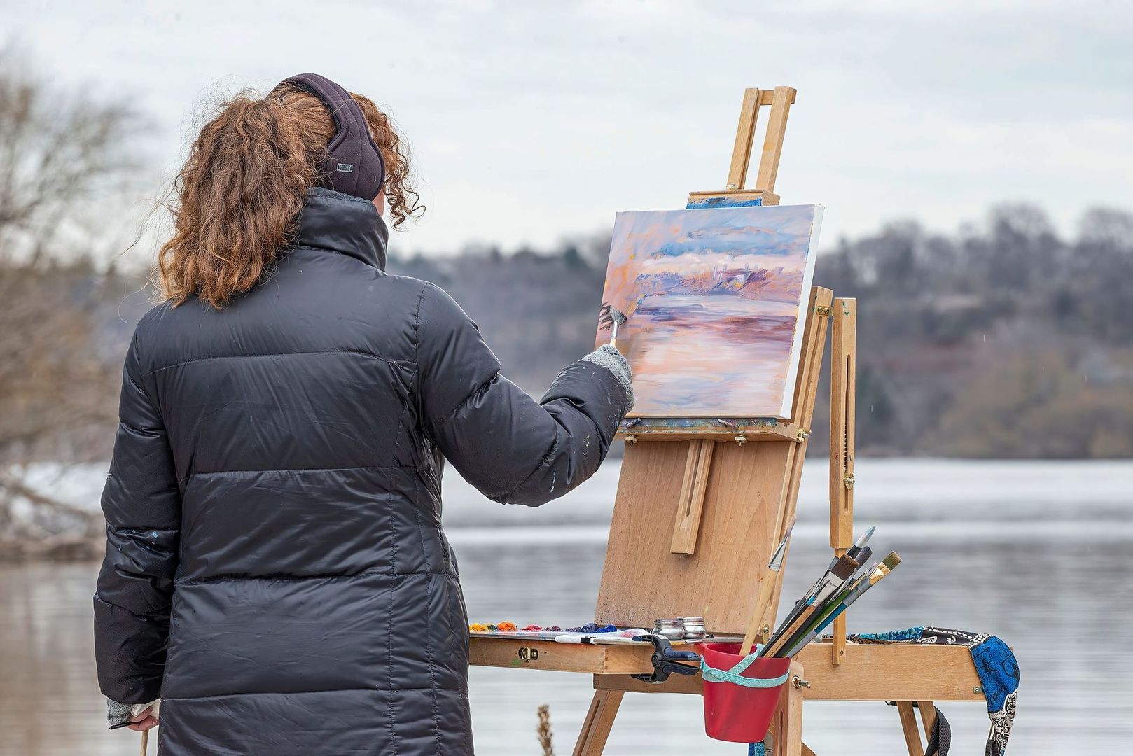 Plein air artist painting a lakeside scene on a wooden easel